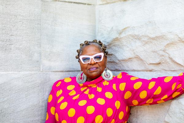Crystal Wilkinson, an award-winning author, former Kentucky Poet Laureate, and University of Kentucky professor of English. She is standing against a stone wall and wearing glasses and a colorful pink and yellow polka dot dress.
